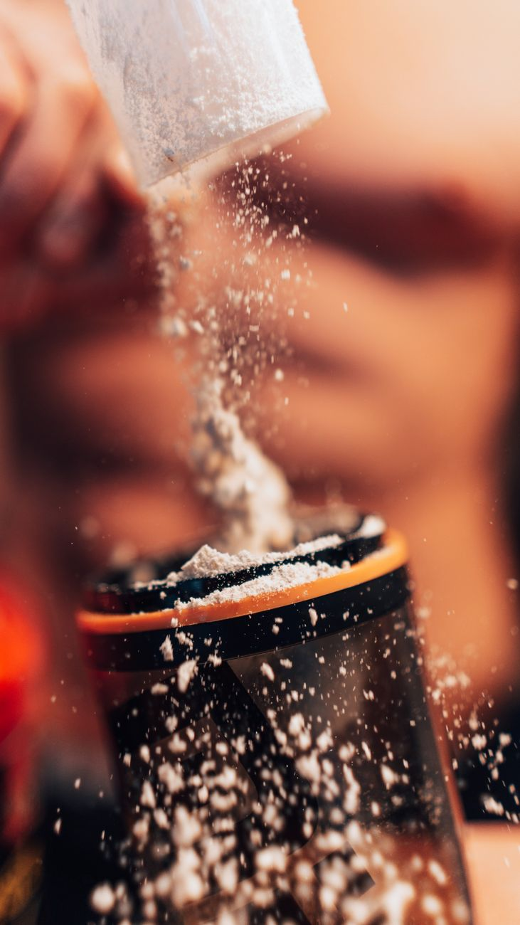 A scoop of white creatine powder on a kitchen bench beside a printed dosing calendar, a shaker bottle, and a smartphone showing a weight graph, illustrating how loading and low-dose protocols reach the same saturation on different timelines