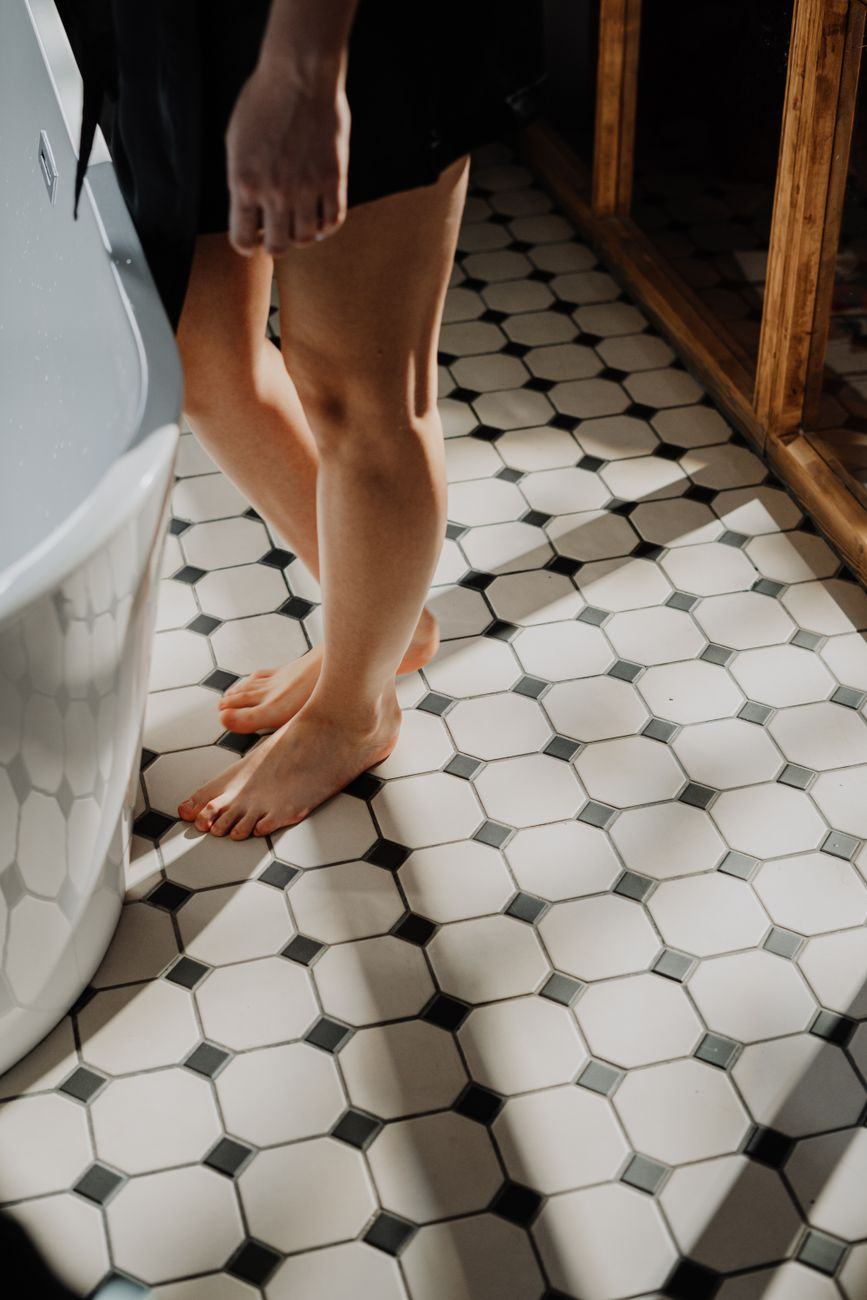 A person standing on a bathroom scale in soft morning light beside a glass of water and a smartwatch on the floor, illustrating the stable step in scale weight that creatine saturation adds and maintenance preserves