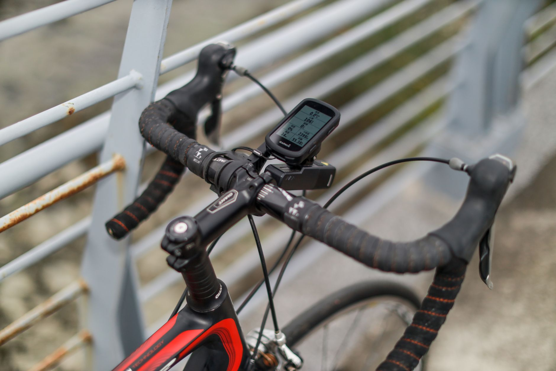 A road cyclist checking a handlebar-mounted bike computer beside a power meter crankset, a heart-rate strap on a bench, and a smartphone showing a cycling calorie graph on a sunlit workshop bench