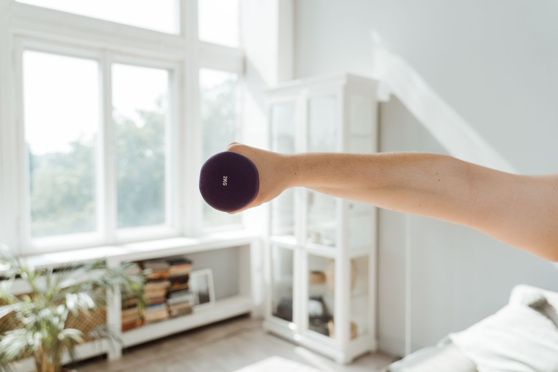 An older adult lifting light dumbbells in a sunlit living room beside a water bottle and a smartphone showing a workout log, illustrating how resistance training preserves muscle through midlife and beyond