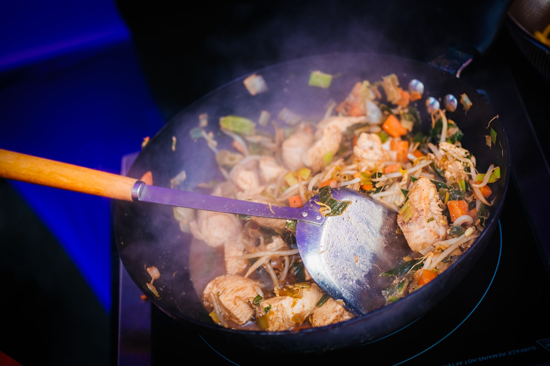 A finished stir-fry portioned on a plate beside the full wok on a scale, a notebook with per-ingredient calorie figures, and a smartphone calorie app on a sunlit kitchen counter, illustrating per-ingredient logging of a homemade meal