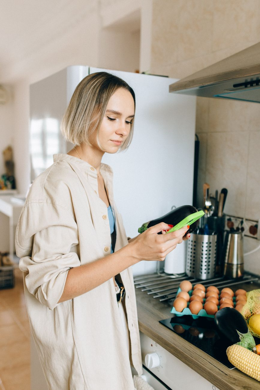 Person logging a meal on a smartphone calorie tracking app at a sunlit kitchen counter beside a digital scale weighing roast vegetables and a small notebook listing food entries