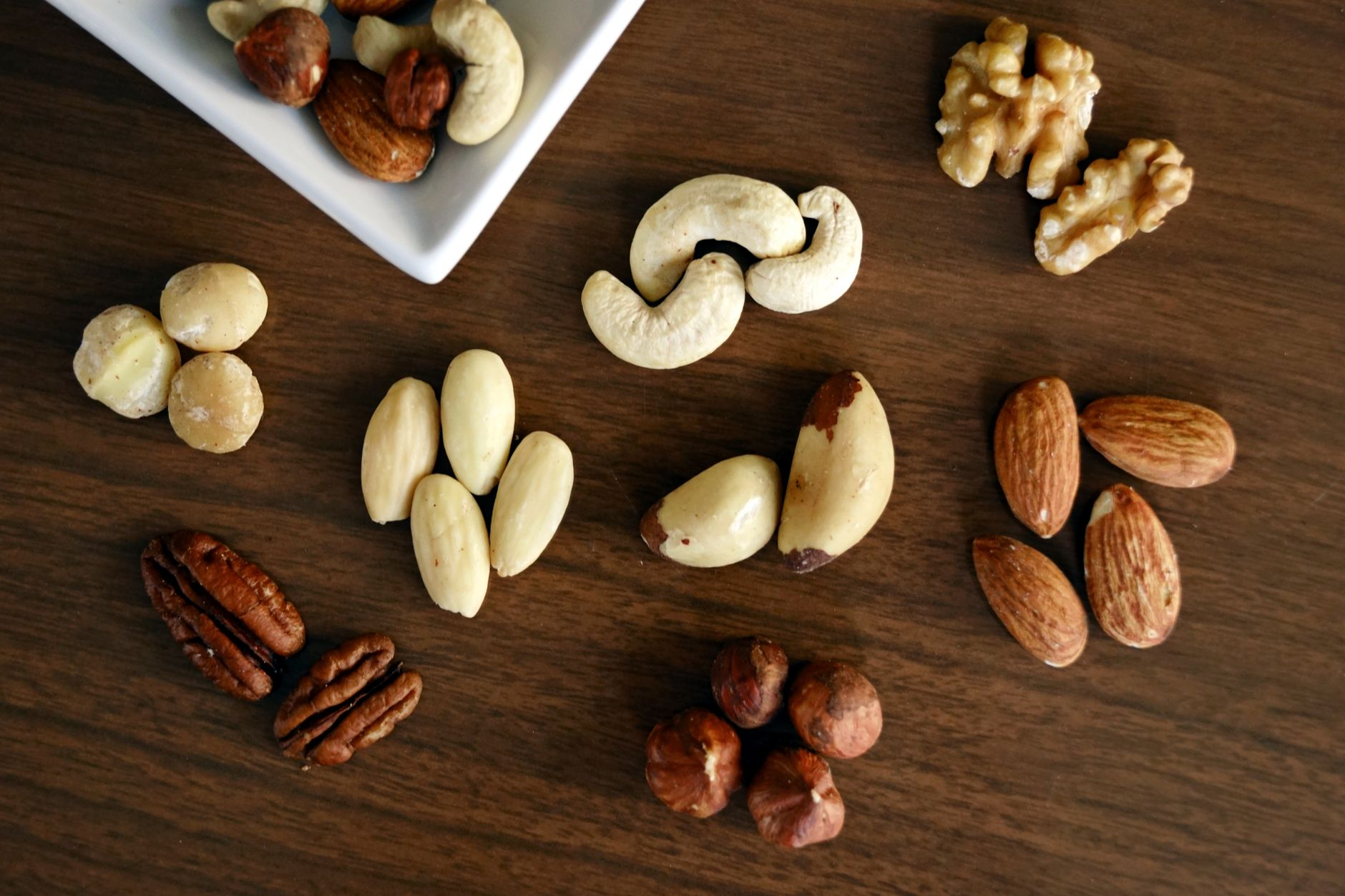 A bowl of mixed nuts — almonds, walnuts, cashews, and pistachios — beside a digital kitchen scale, a printed nutrition label, and a smartphone showing a calorie tracking app on a sunlit kitchen counter, illustrating the gap between labelled and absorbed nut calories