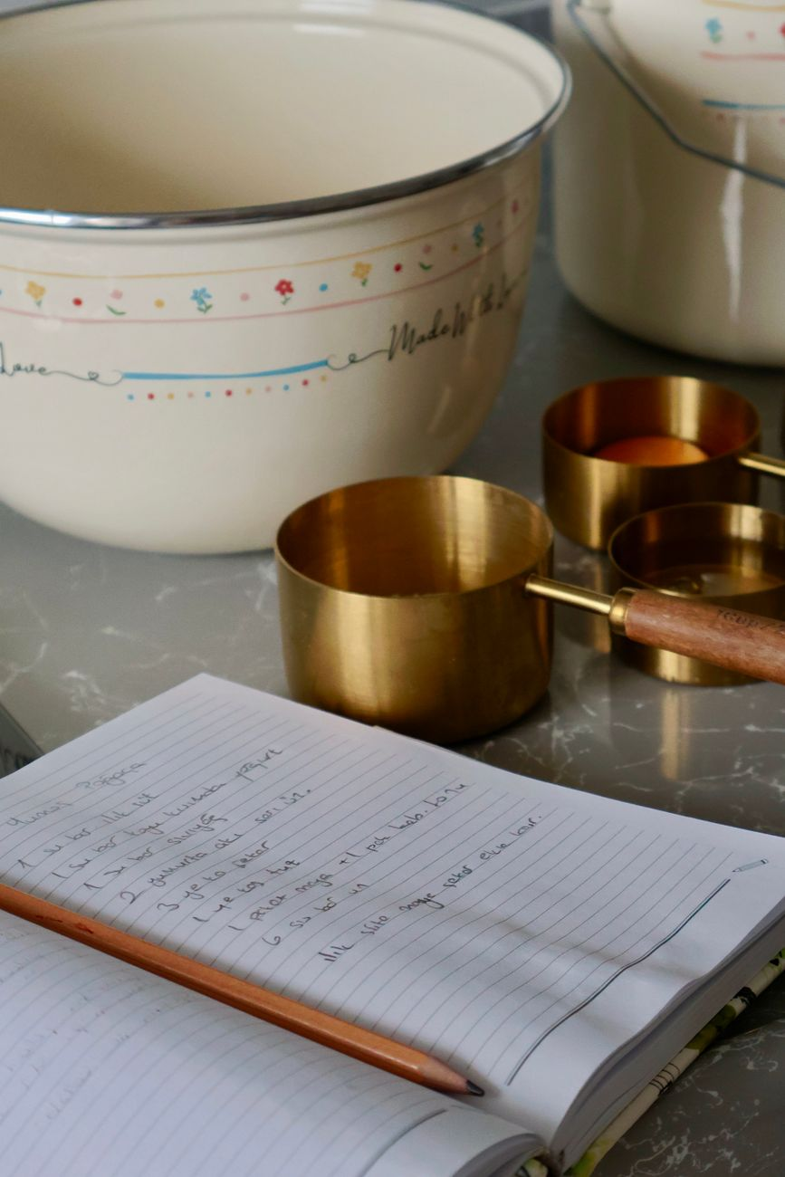 An open notebook with a handwritten raw-to-cooked conversion table beside a bowl of cooked rice, a kitchen scale, and a smartphone calorie app on a sunlit kitchen counter
