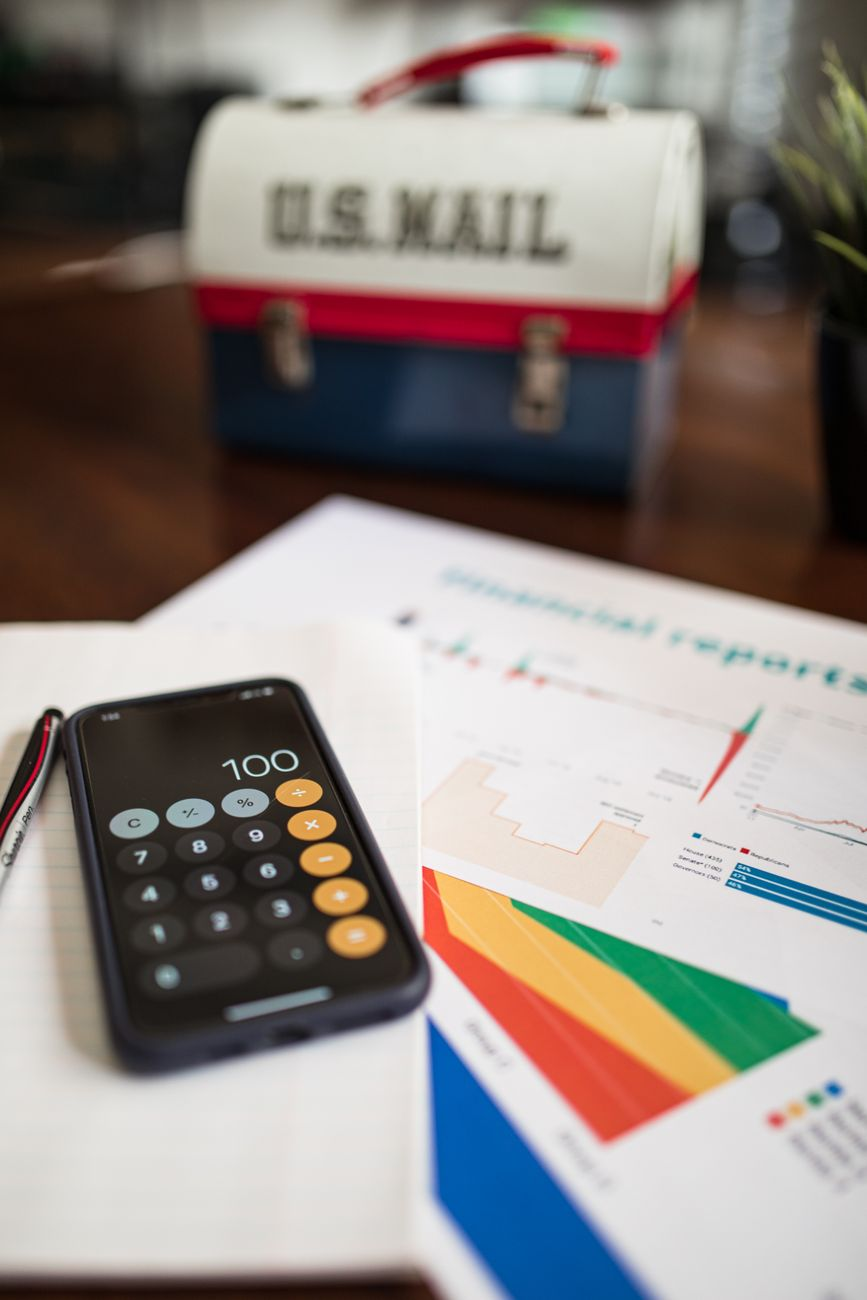 A loaded rucksack on a kitchen scale beside a printed calorie table, a calculator, and a smartphone showing a tracking app on a sunlit counter, illustrating how pack weight maps to calories per hour