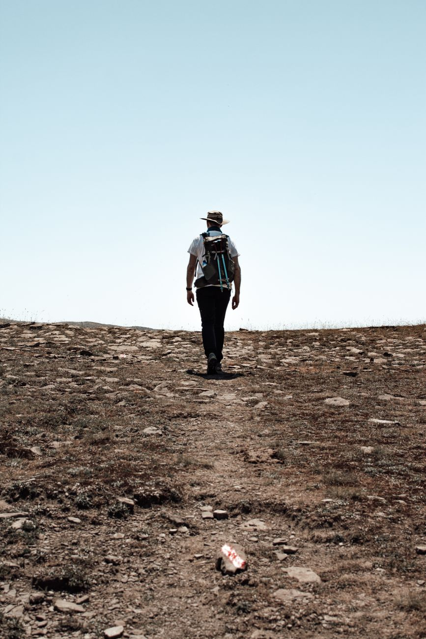 A hiker with a loaded backpack walking up a sunlit hill trail, illustrating how grade and load together drive rucking calorie burn far more than the pack alone