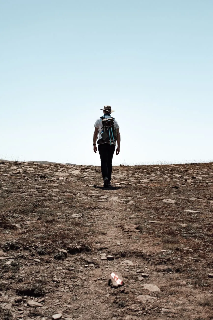 A hiker with a loaded backpack walking up a sunlit hill trail, illustrating how grade and load together drive rucking calorie burn far more than the pack alone