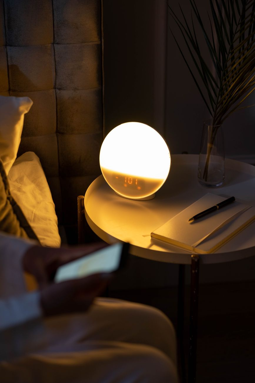 A bedside table at dawn with an alarm clock reading 5 a.m., an open notebook with sleep-hour tally marks, a smartphone showing a sleep-tracking graph, and a glass of water on a sunlit windowsill