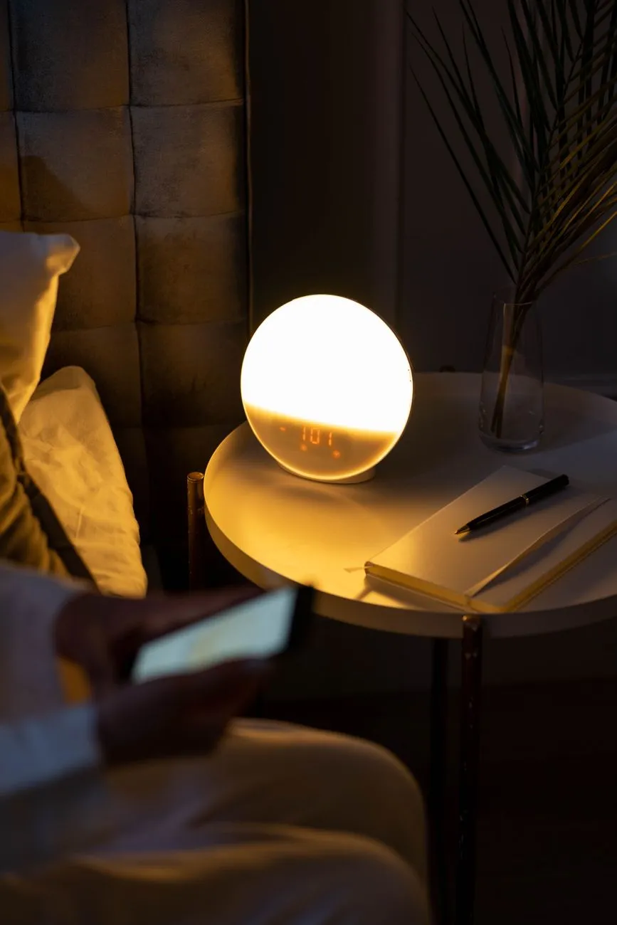 A bedside table at dawn with an alarm clock reading 5 a.m., an open notebook with sleep-hour tally marks, a smartphone showing a sleep-tracking graph, and a glass of water on a sunlit windowsill