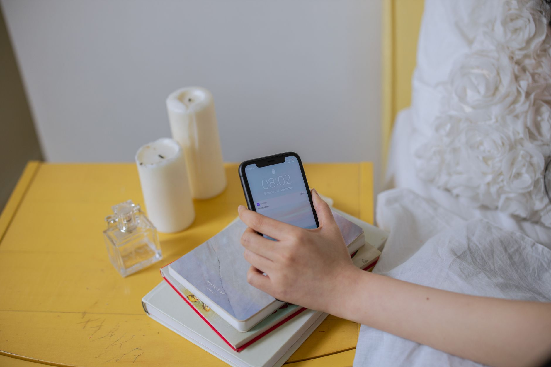 A bedside table at dawn with an alarm clock reading 5 a.m., an open notebook with sleep-hour tally marks, a smartphone showing a sleep-tracking graph, and a glass of water on a sunlit windowsill