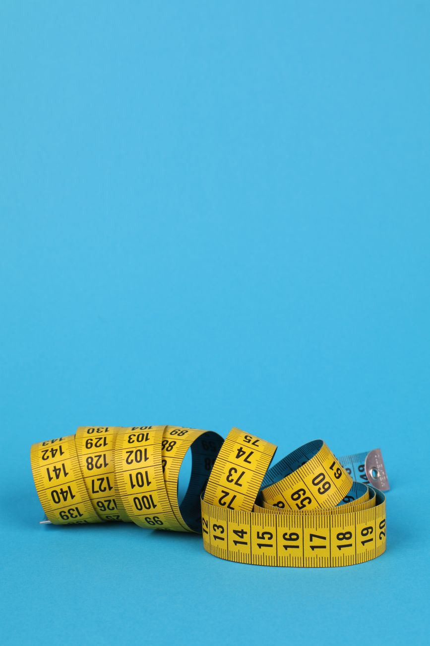 A smart bathroom scale beside a printed table comparing body fat readings from consumer scales with DXA results, a calculator, and a tape measure on a sunlit bathroom floor, illustrating the per-device error ranges found in validation studies