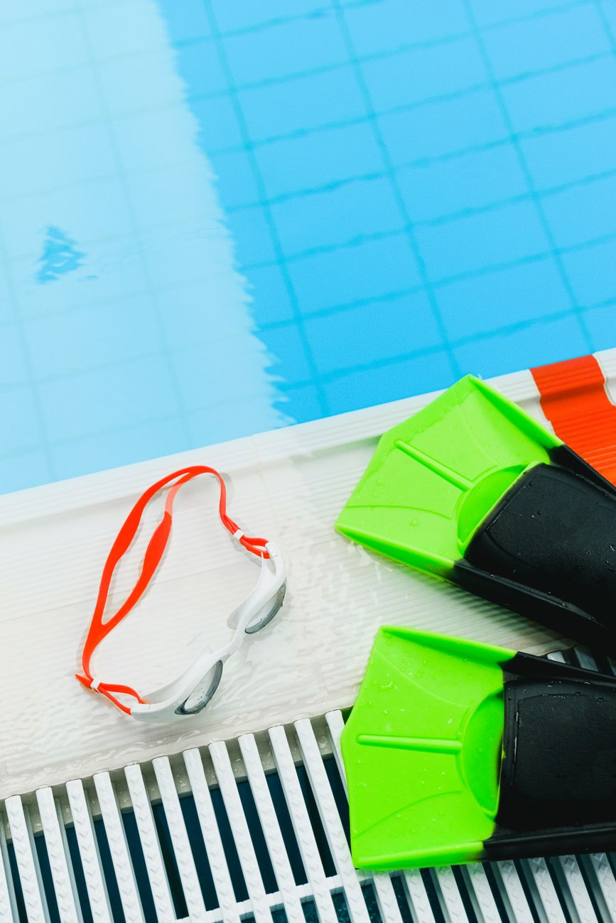 A printed table of swimming MET values by stroke beside a calculator, swim goggles, and a smartphone showing a calorie tracking app on a sunlit pool deck, illustrating the stroke-by-stroke math behind swimming calorie burn