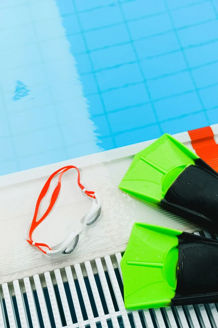 A printed table of swimming MET values by stroke beside a calculator, swim goggles, and a smartphone showing a calorie tracking app on a sunlit pool deck, illustrating the stroke-by-stroke math behind swimming calorie burn