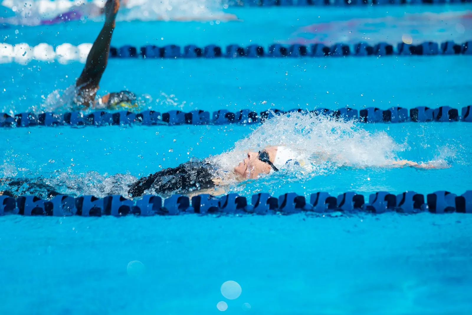A swimmer doing freestyle laps in a sunlit pool beside a smartwatch showing a calorie-burn number and a smartphone with a calorie tracking app on the pool edge, illustrating how stroke, pace, and body weight set swimming calorie burn